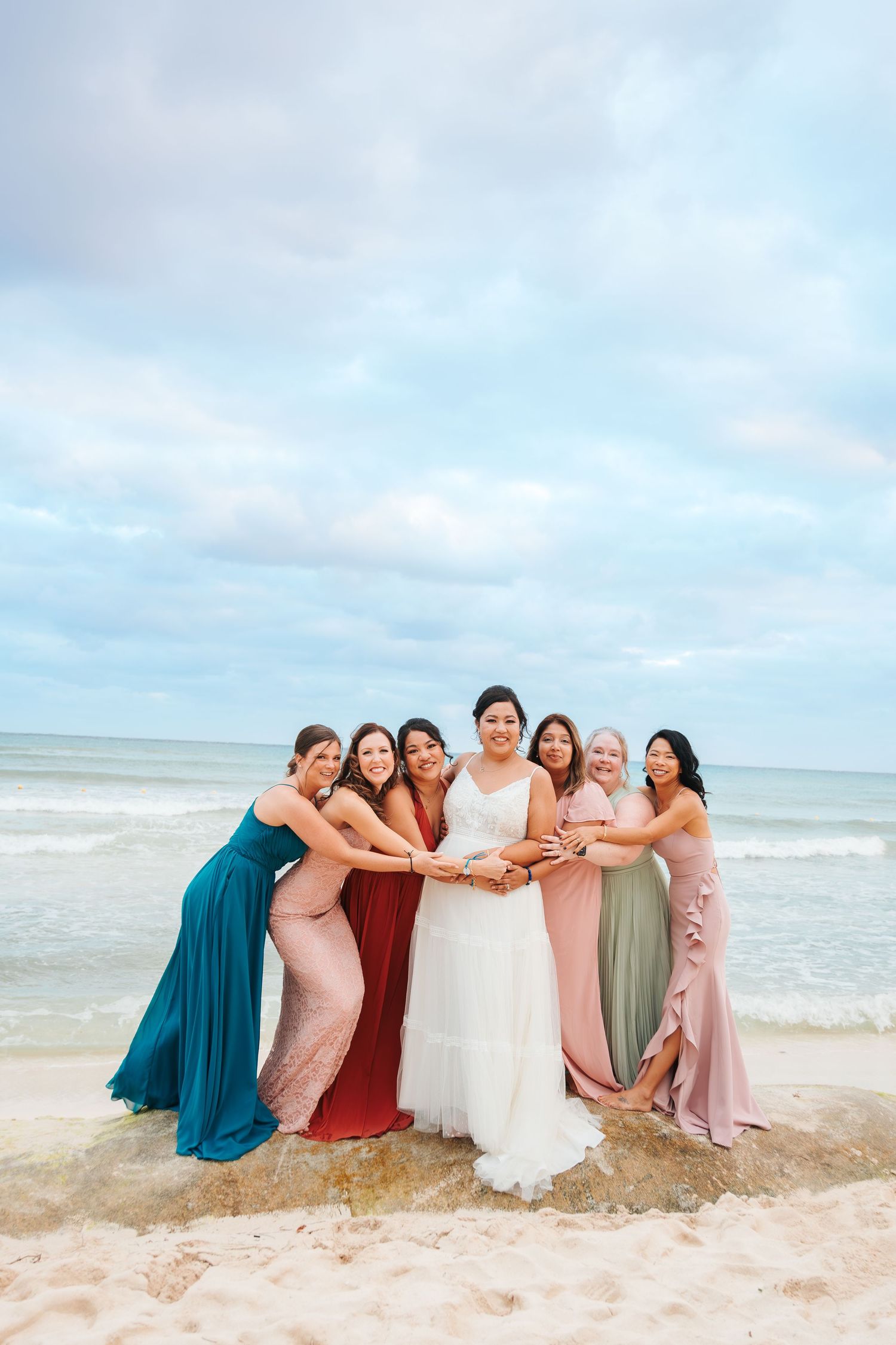 Bride in white gown poses with six bridesmaids in colorful dresses on a sandy beach with ocean waves behind them. destination wedding