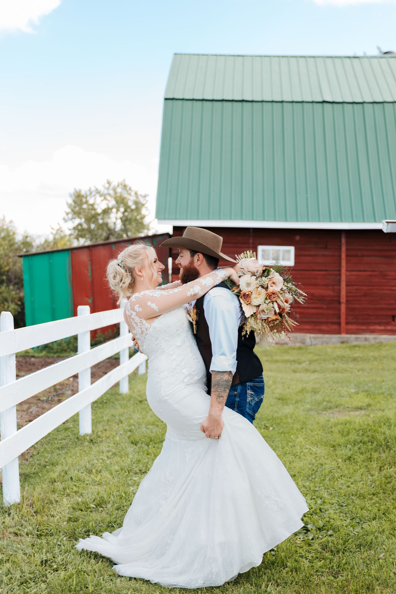 Bride and groom embracing in front of a rustic red barn with a green roof on a sunny day. western weddings. Banff elopement pictures