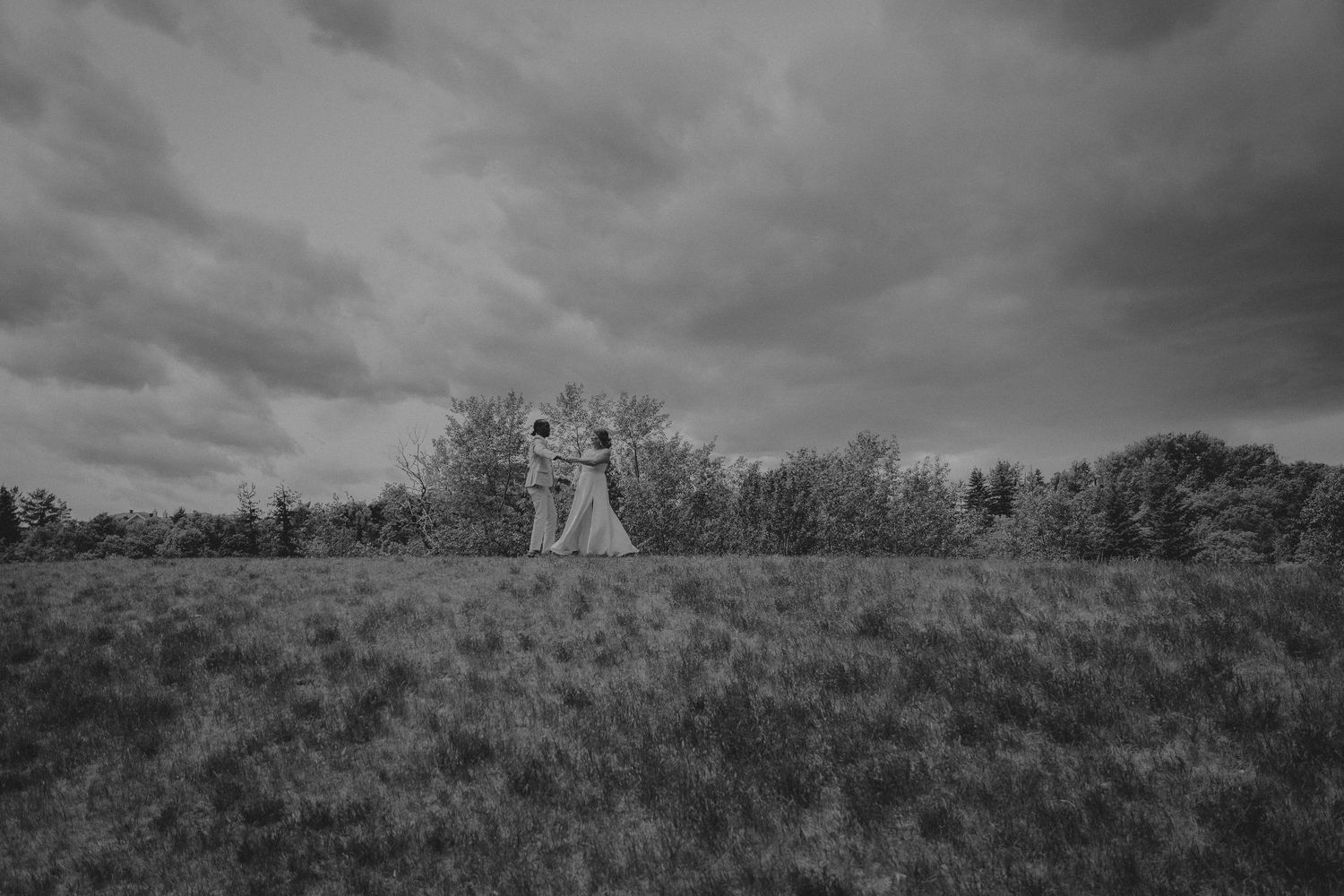 Black and white photo of a couple dancing in a vast grassy field under dramatic cloudy skies. Elopement Pictures. Destination weddings.