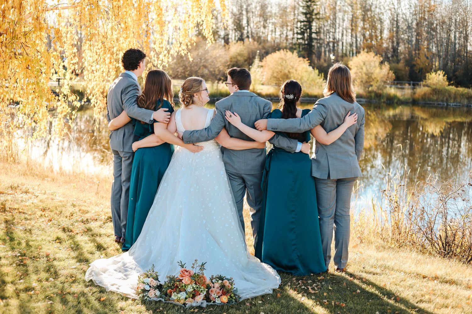 Wedding party in gray and teal standing arm-in-arm by a sunlit autumn pond, bride in white gown centered. Fall wedding pictures outside.