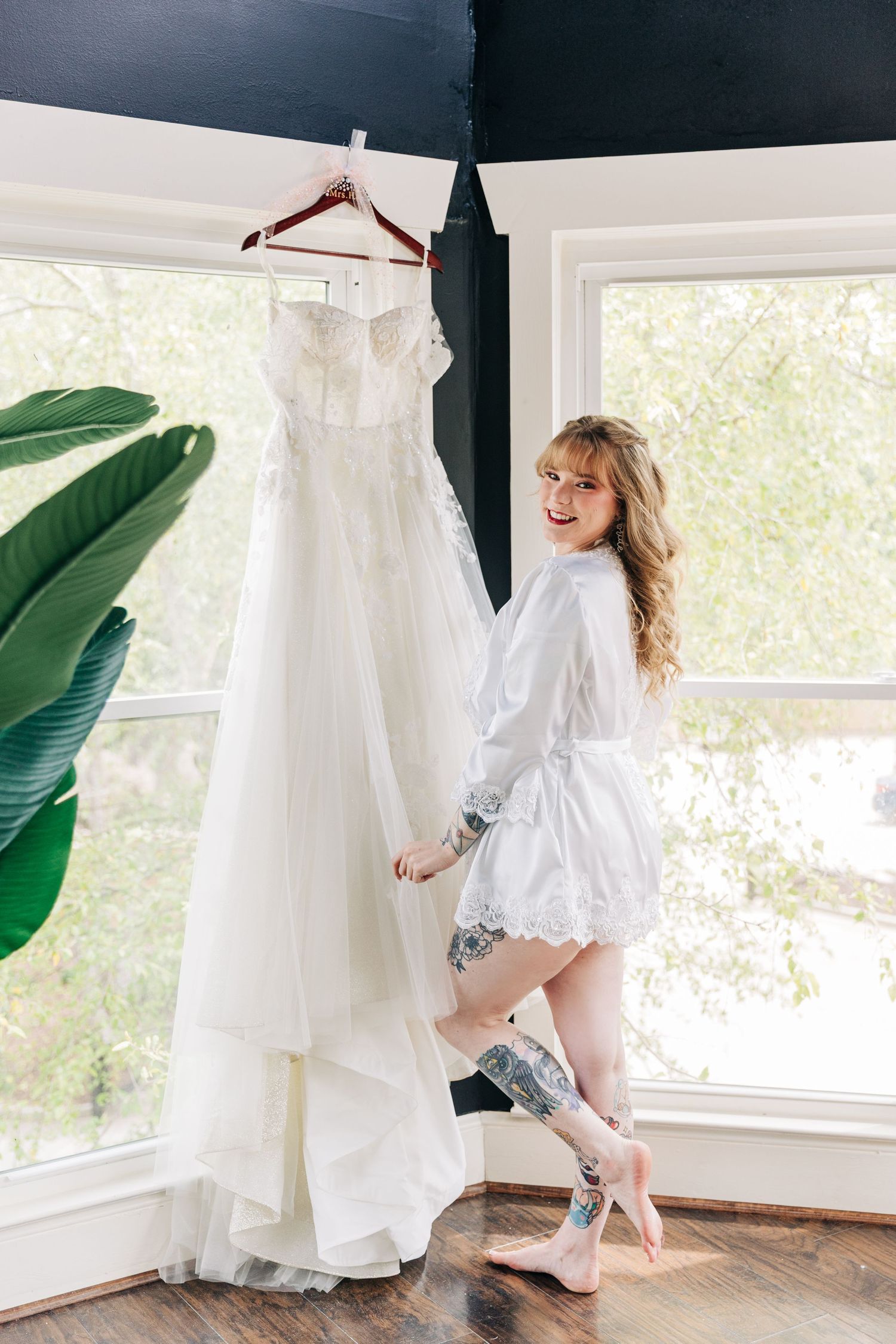 Smiling bride in white lace robe stands beside her elegant strapless wedding gown hanging by a sunlit window.