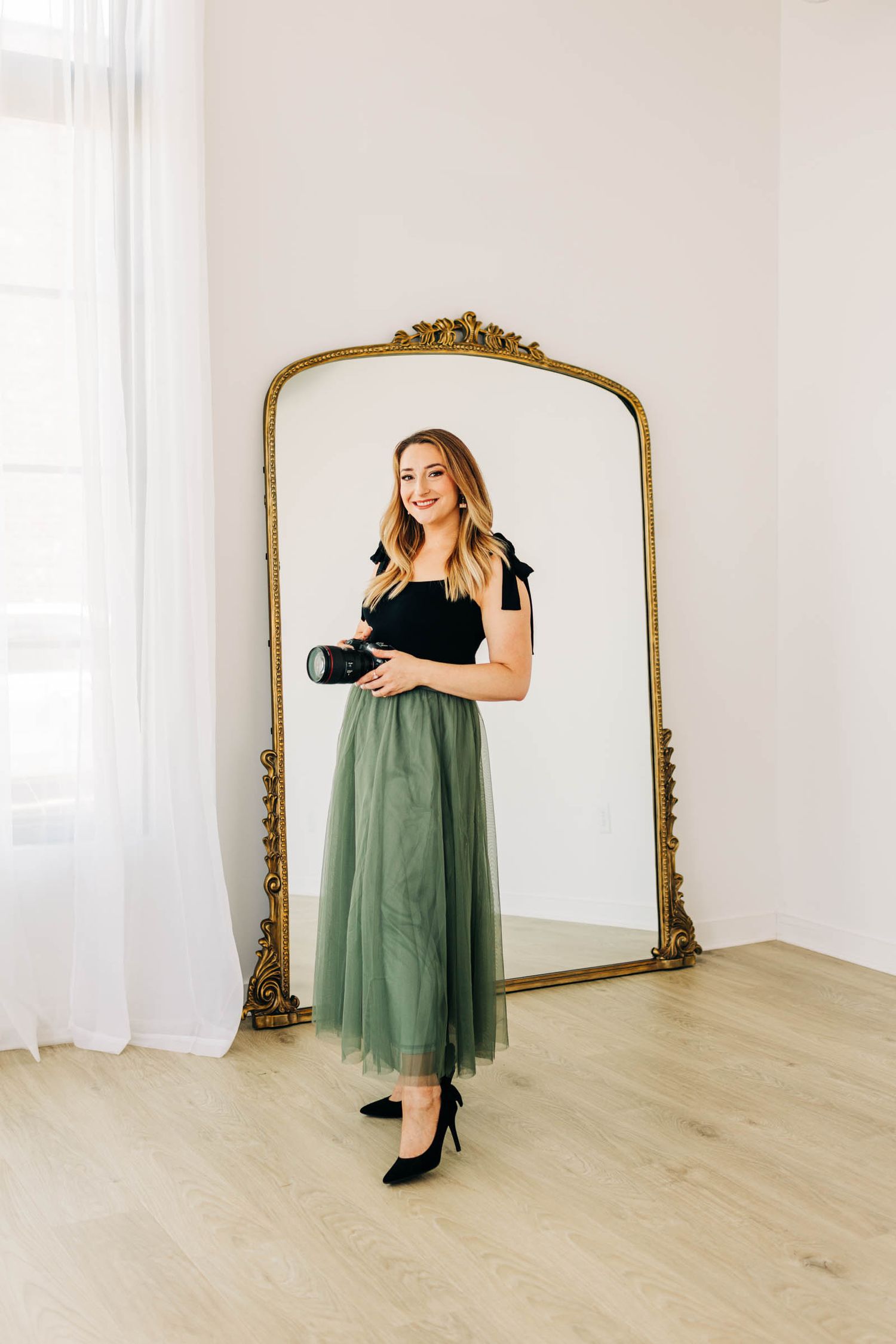 Woman in green tulle skirt holding camera stands before ornate gold mirror in bright white studio.
