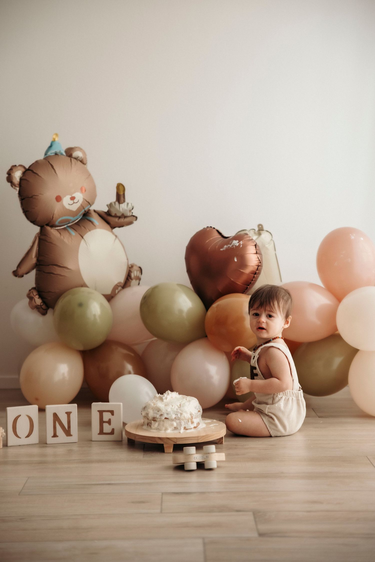 Baby's first birthday cake smash with bear balloon, colorful balloons, and ONE blocks on wood floor.