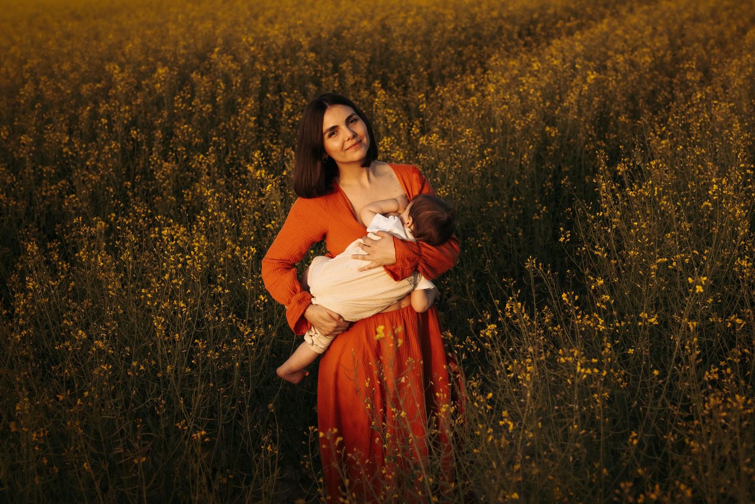 A person in a flowing red dress stands in a golden wildflower field at sunset holding a small child.