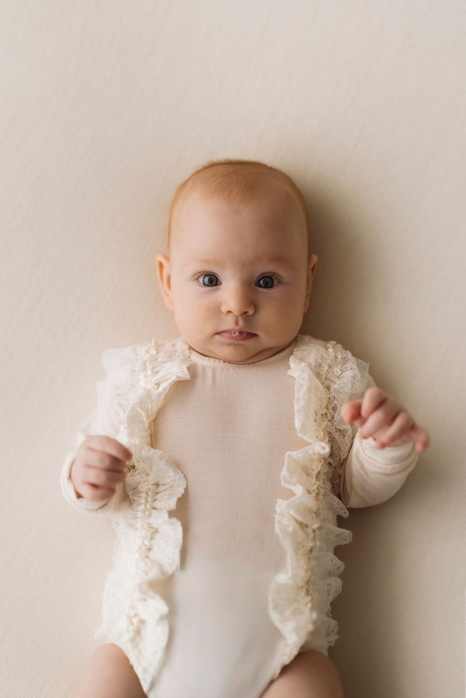 Baby in white ruffled outfit lying on beige background in artistic minimalist style portrait series.