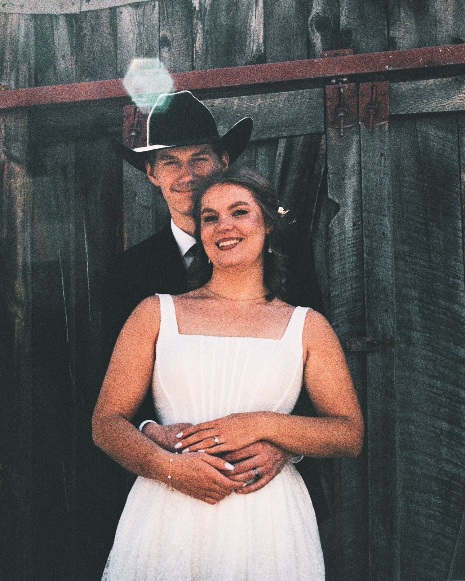 Couple in wedding attire posing by rustic barn, groom in black cowboy hat embracing bride in white dress.