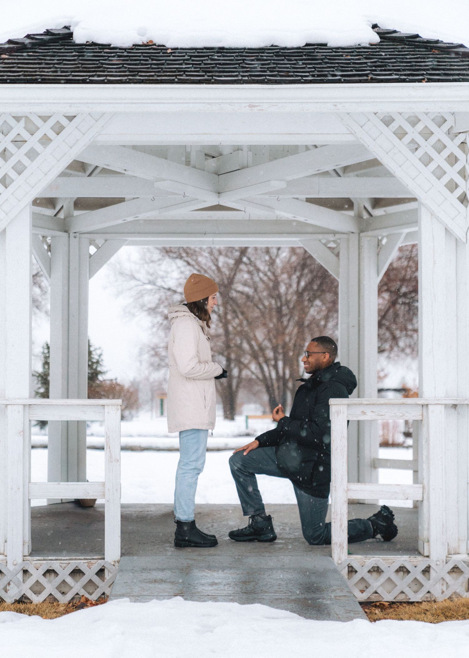 Man proposing on one knee to his partner inside a white gazebo surrounded by snow in winter.