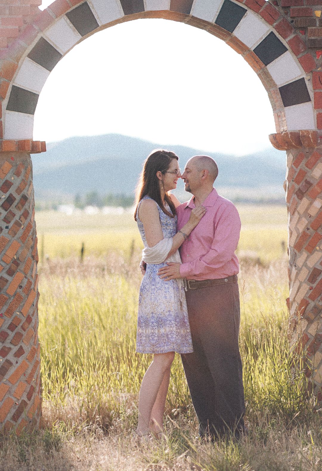 Romantic couple nearly kissing under a decorative brick arch framed by a golden countryside backdrop.