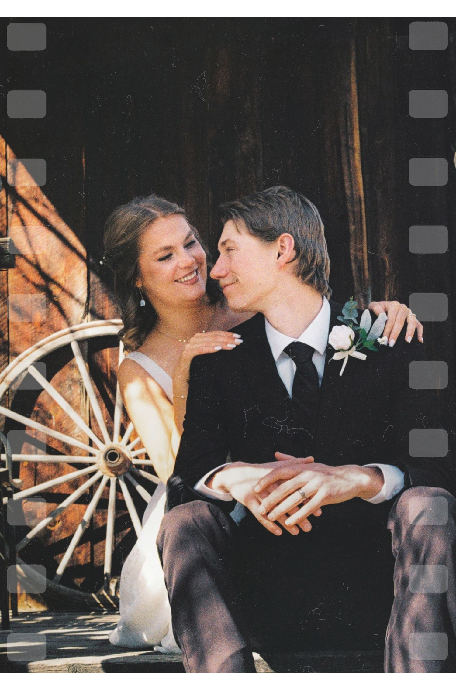 A newlywed couple in wedding attire sits together smiling beside a rustic wooden barn and vintage wagon wheel.