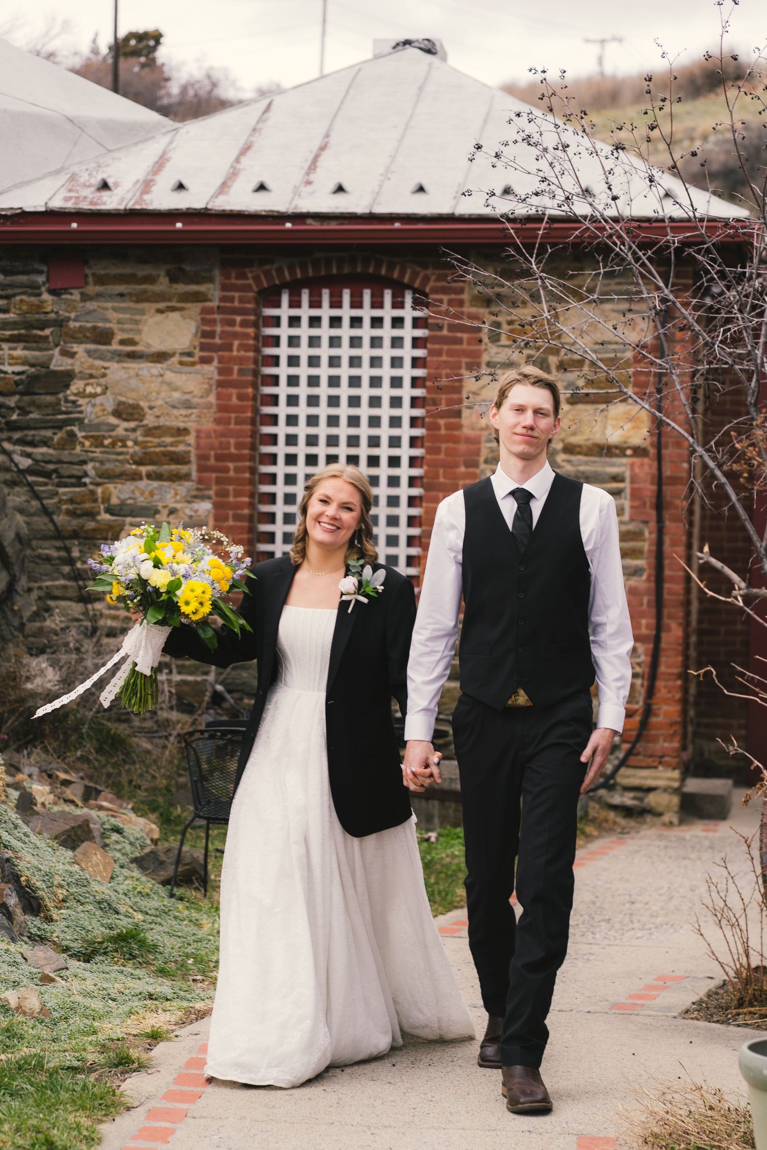 Bride and groom walking hand in hand holding yellow bouquet outside rustic brick building.