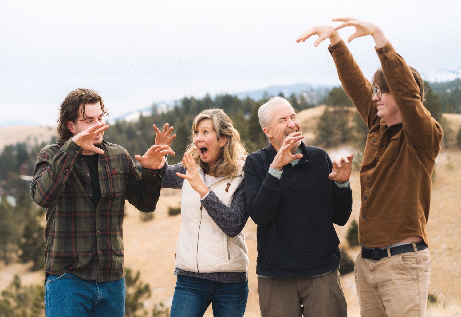 Family of four laughing and making fun poses outdoors in a scenic mountain setting.