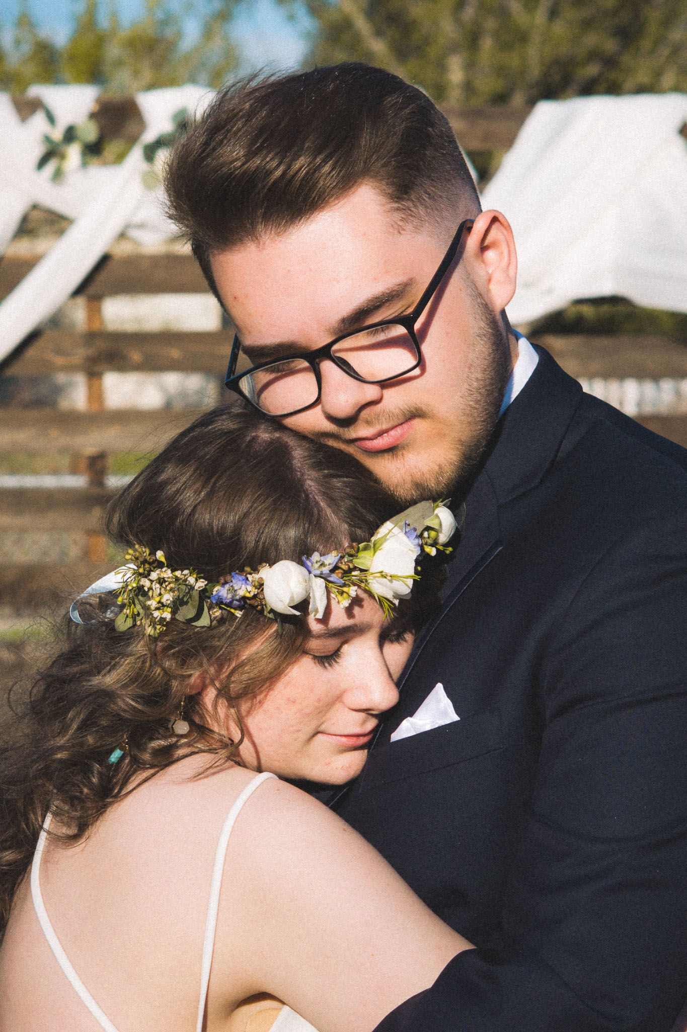 A groom in glasses and black suit embraces his bride wearing a floral crown at an outdoor wedding ceremony.