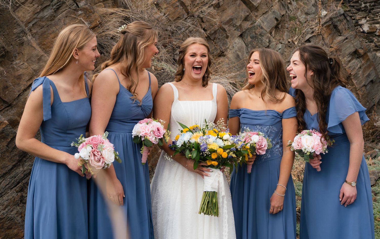 Bride and four bridesmaids in blue dresses laughing joyfully while holding colorful bouquets outdoors.