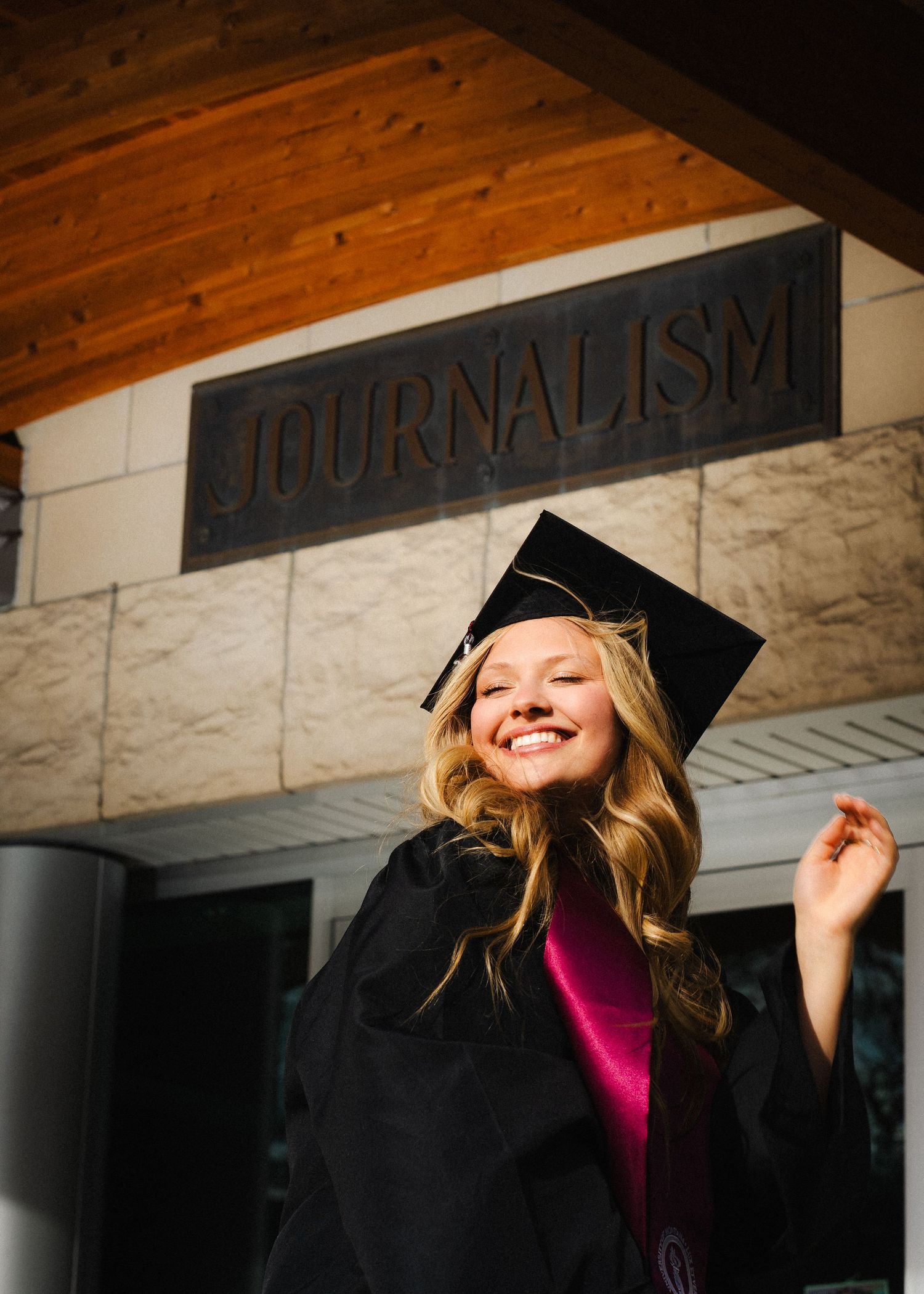 Smiling female graduate in black cap and gown poses in front of a Journalism building sign on graduation day.