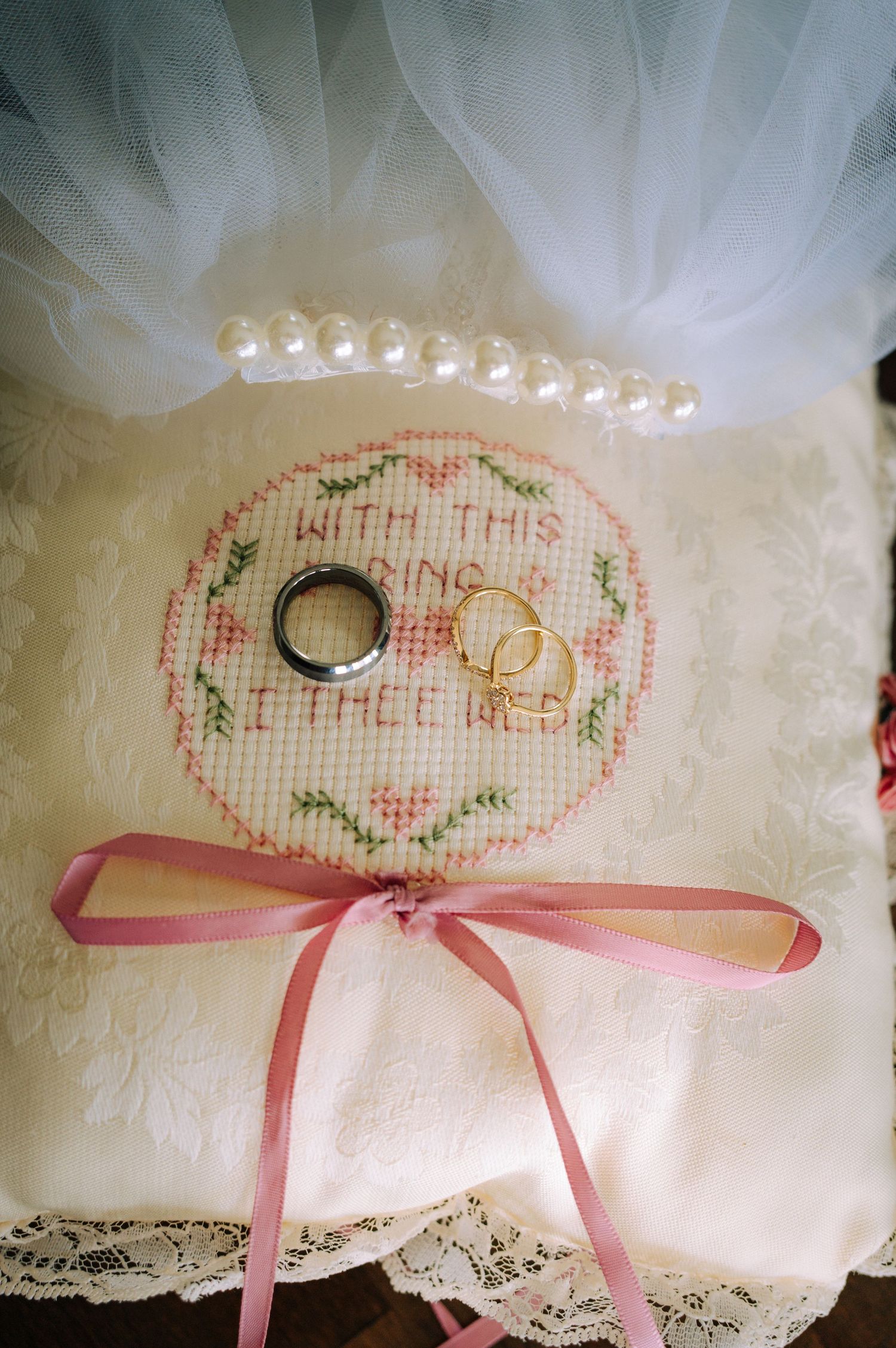 Cross-stitch ring bearer pillow with wedding rings and pearl bracelet, pink ribbon bow, reads With This Ring I Thee Wed.
