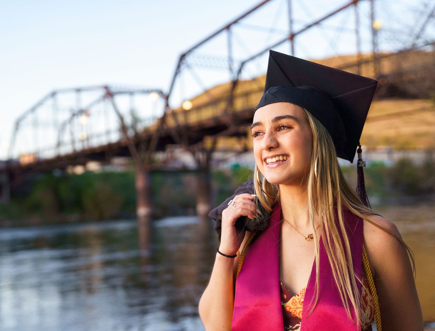 A smiling graduate wearing a black cap holds her gown near a metal bridge over a river at sunset.