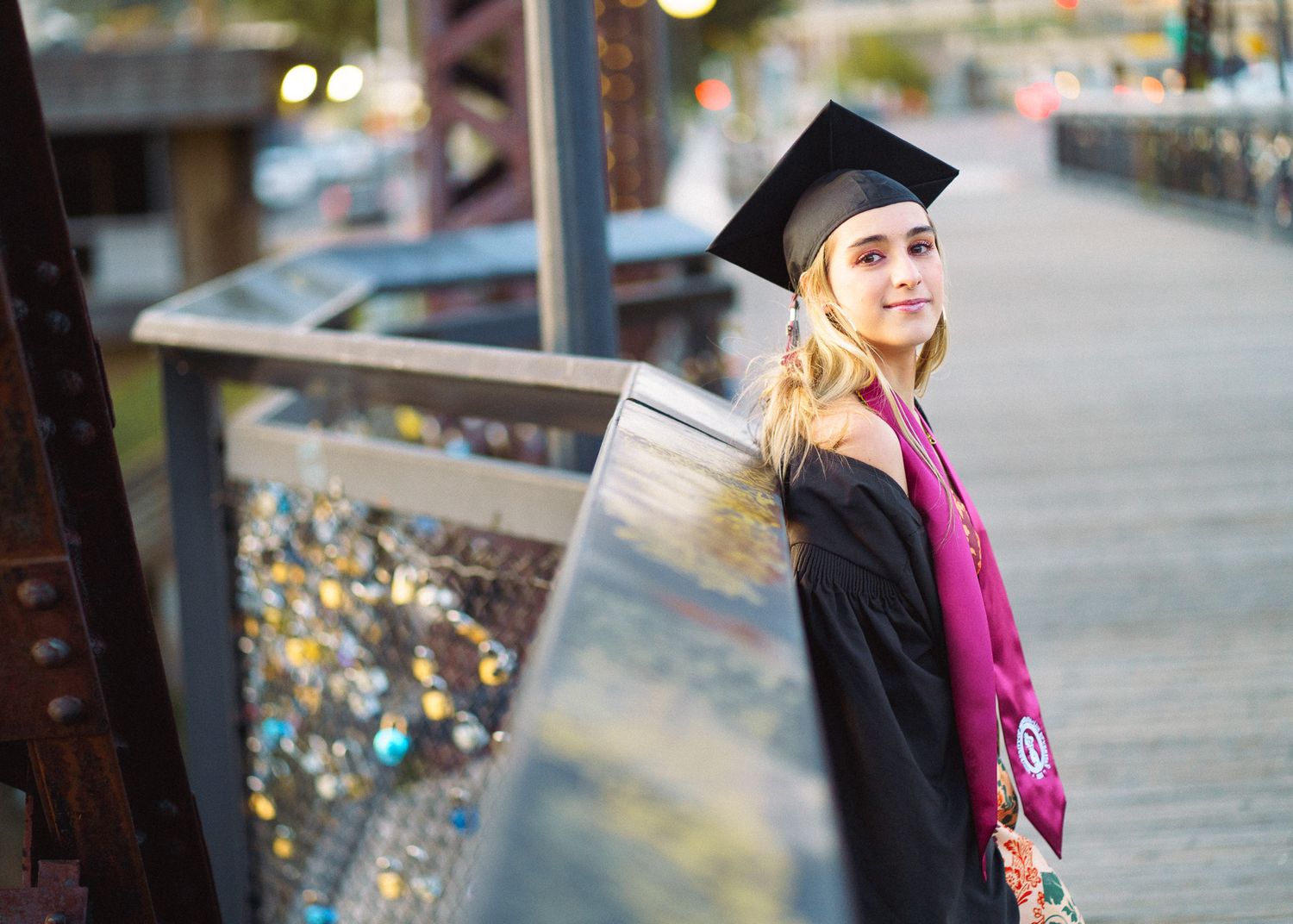 Smiling graduate in black cap and gown with purple stole poses on a bridge during outdoor graduation photo session.