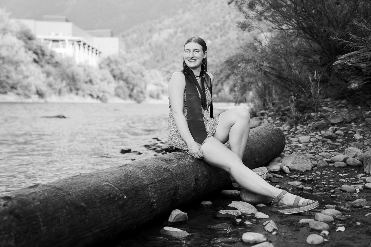 Black and white senior portrait of smiling girl sitting on a log by a rocky riverside.