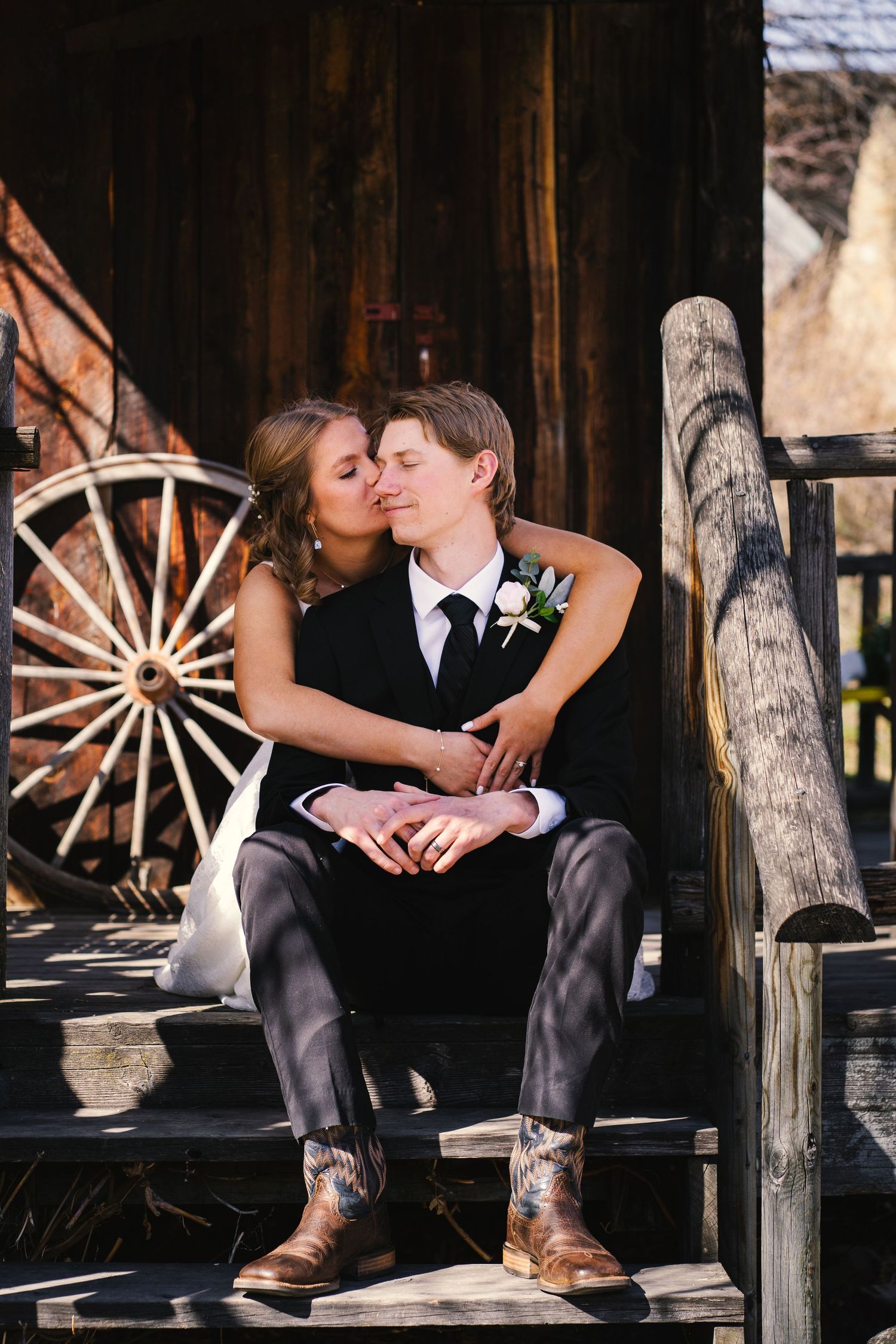 Bride kisses groom on cheek as they sit on wooden steps with wagon wheel backdrop.