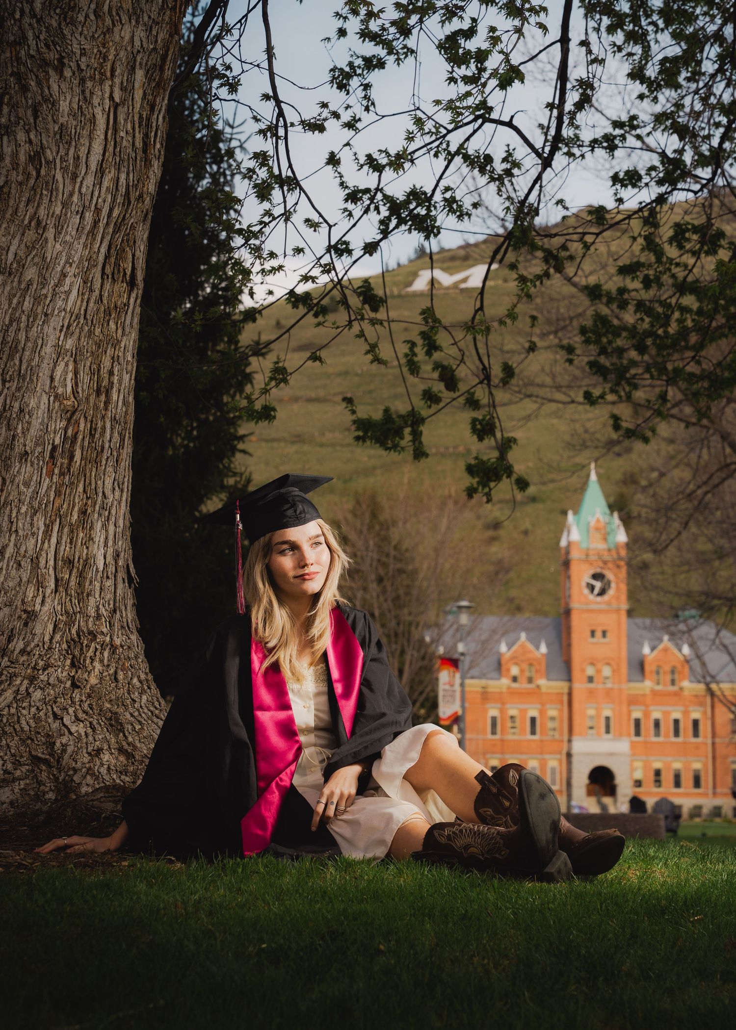 Young woman in graduation cap and gown leaning against tree on Missoula campus lawn at golden hour. Best senior photos Missoula Montana