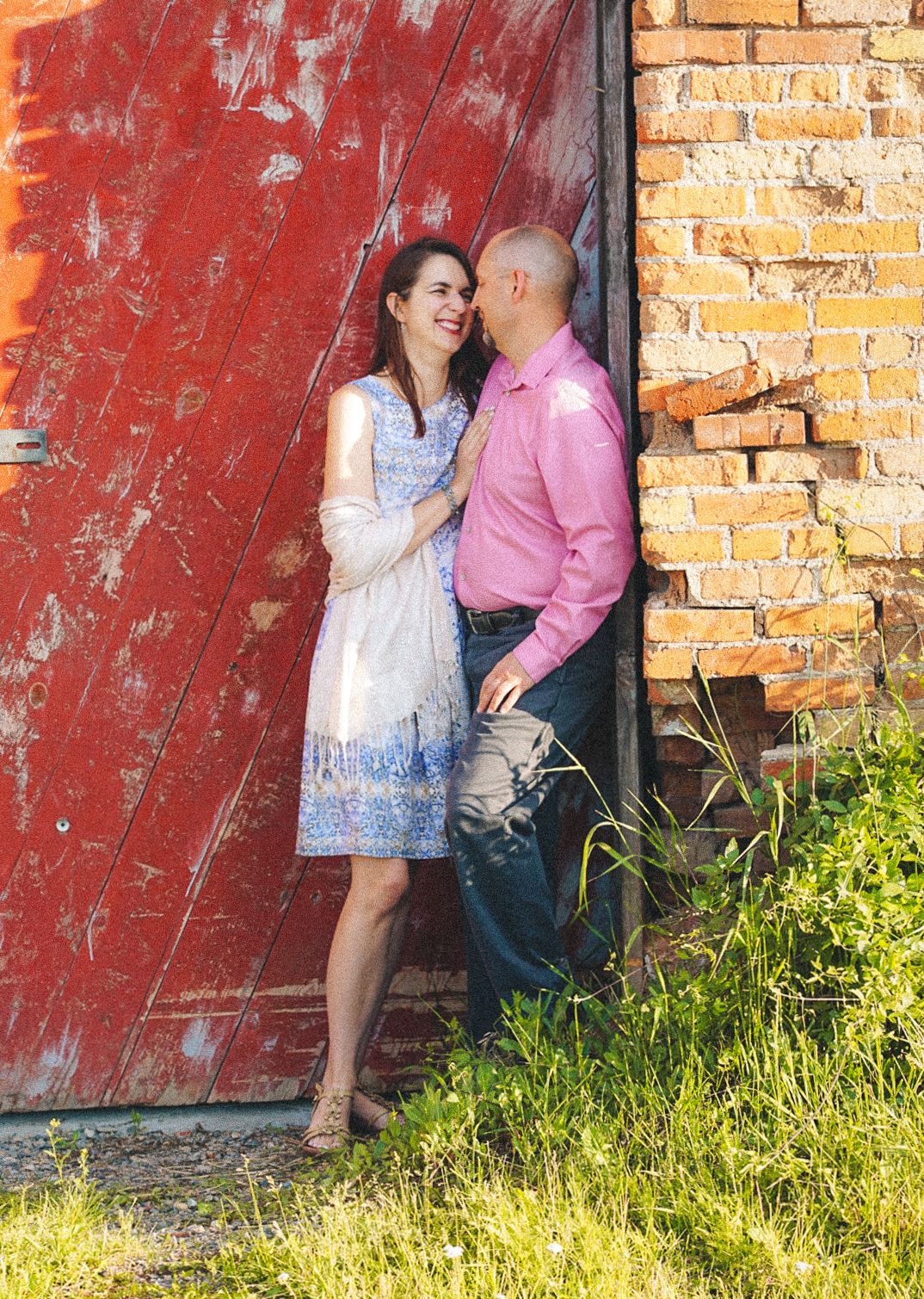 Couple smiling at each other leaning against a rustic red barn door with brick wall backdrop outdoors.