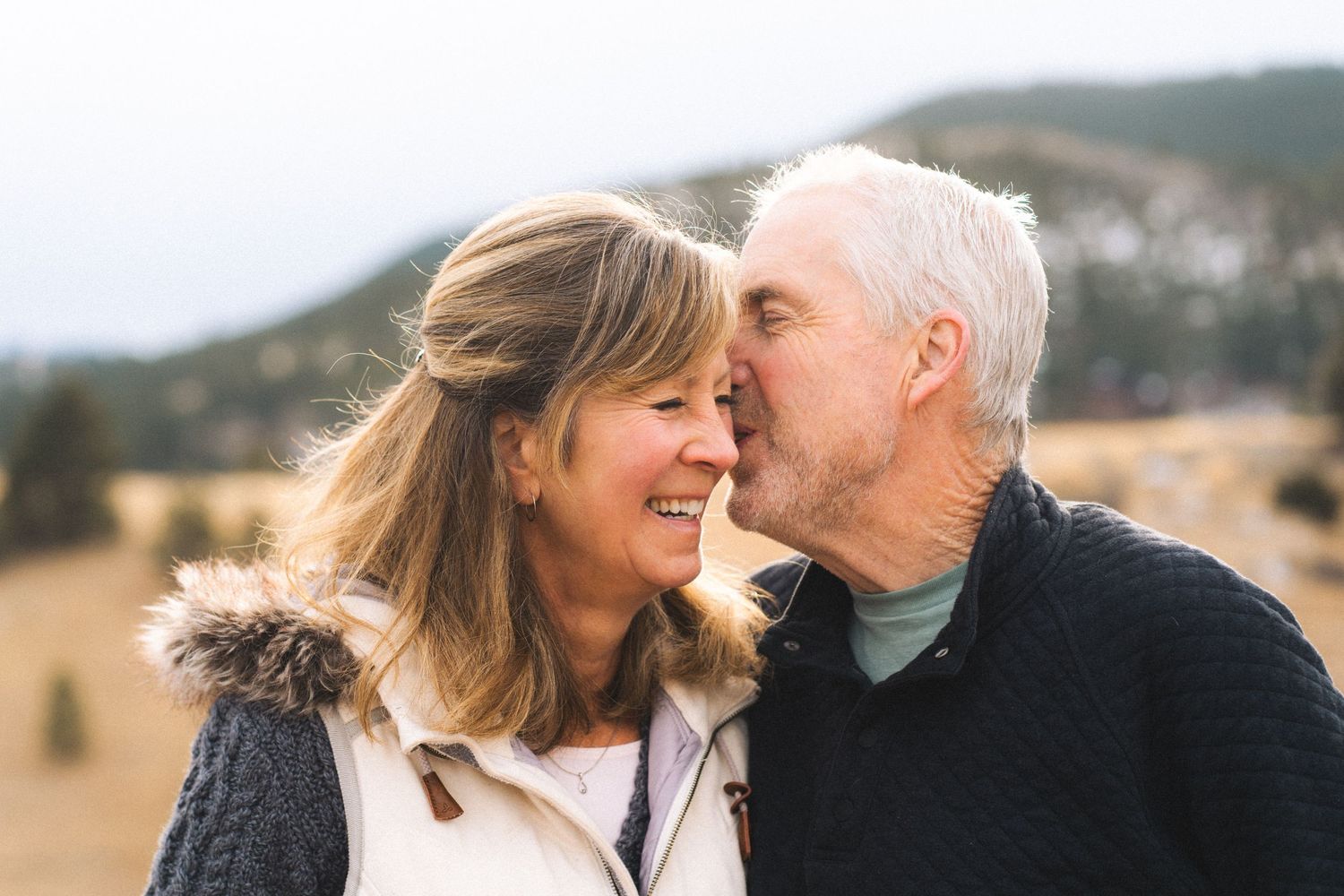 Older couple sharing a sweet kiss outdoors with blurred mountain landscape in the background.