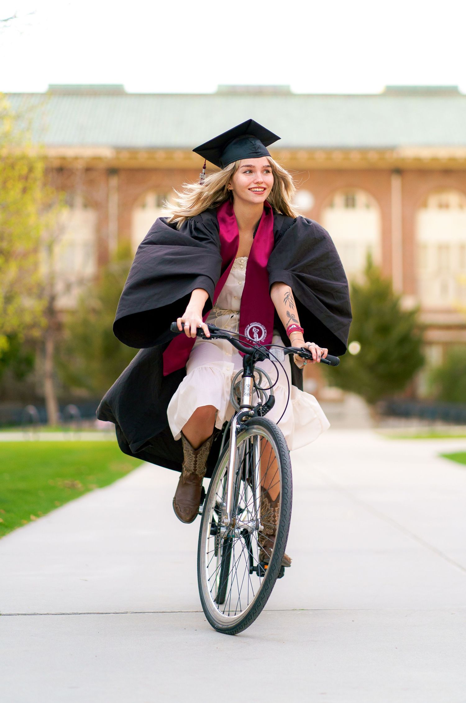 Joyful female graduate in cap and gown riding a bicycle on a tree-lined campus pathway.