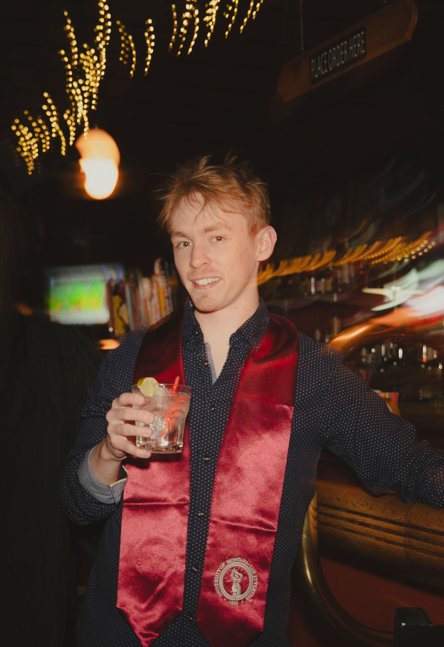 Young man in dark shirt with red velvet graduation sash holds a cocktail at a dimly lit bar.