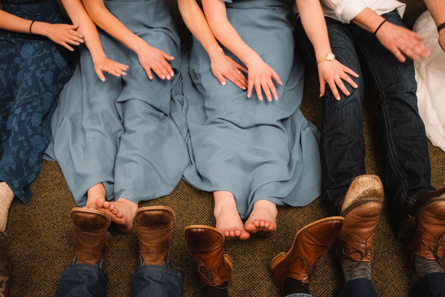 Bridesmaids in blue dresses and groomsmen in brown boots lying on the floor showing hands and feet.