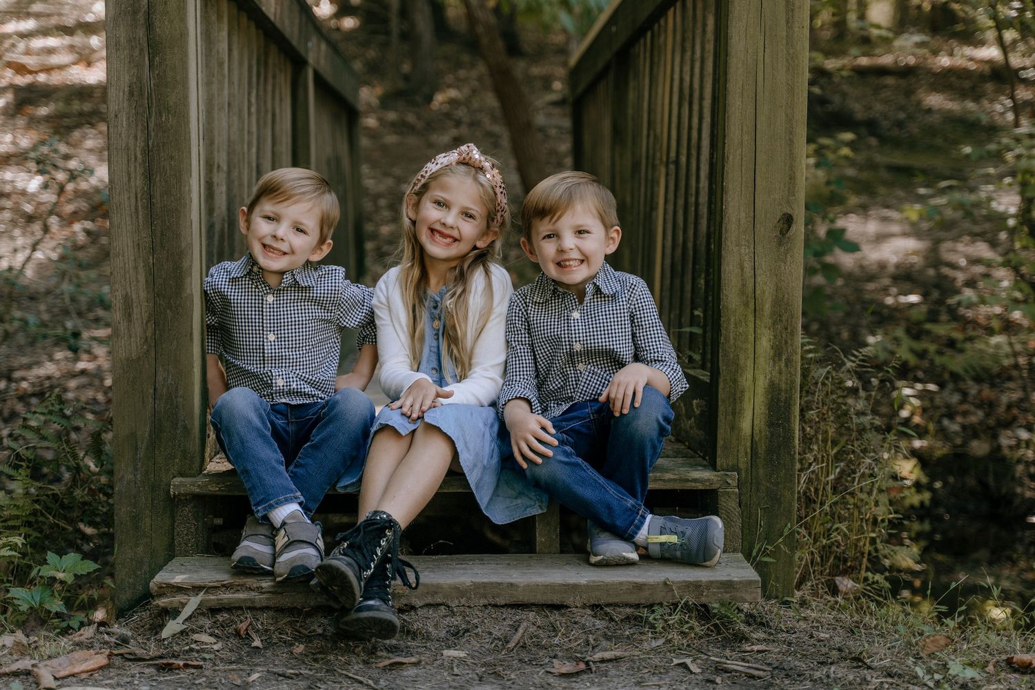 Outdoor portrait of three smiling children sitting together on wooden steps in a rustic forest setting.