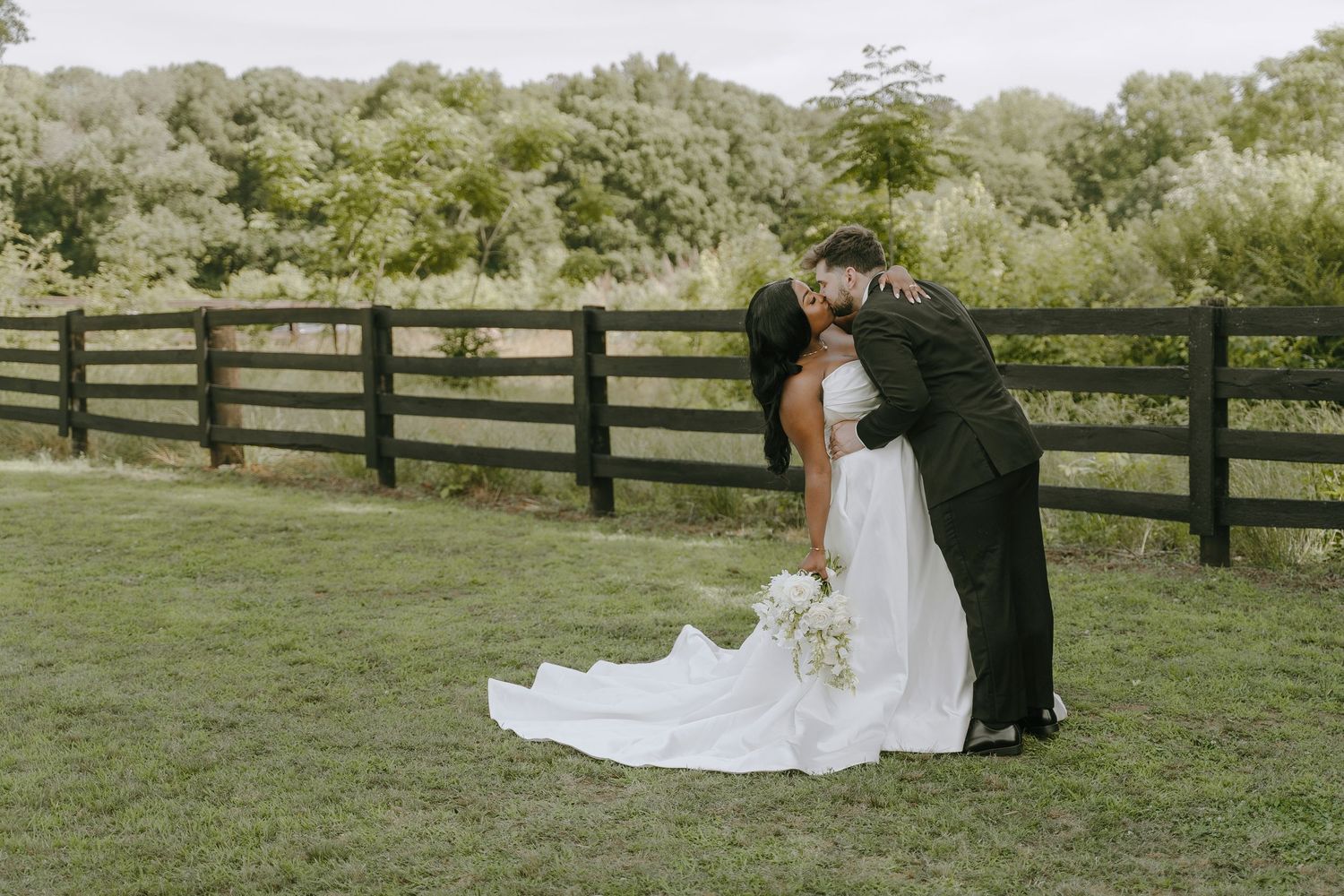 Romantic outdoor wedding photo of a groom dipping and kissing his bride in front of a rustic wooden fence and greenery.