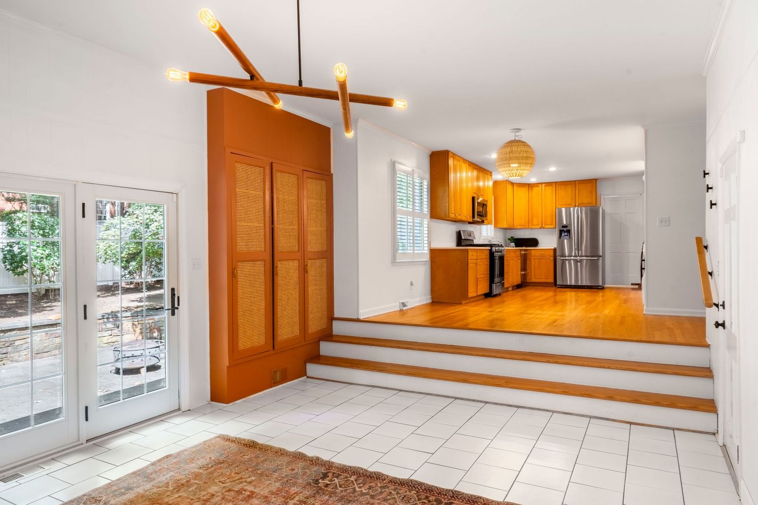Modern open kitchen with warm wood cabinets, stainless steel appliances, and a split-level layout with white tile flooring and French doors.