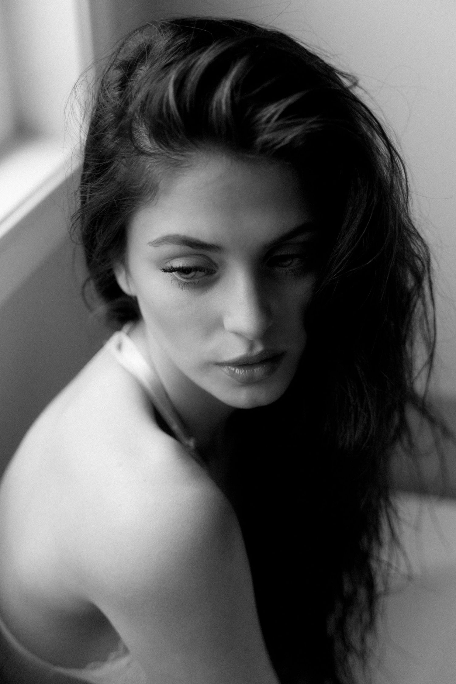 Black and white sultry portrait of a woman with long hair, gazing away thoughtfully by a window.