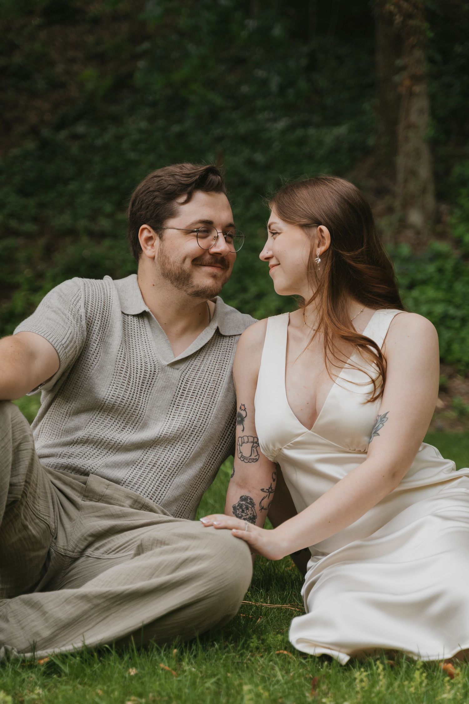 Romantic outdoor portrait of a couple sitting on grass, holding hands, and smiling at each other in soft natural light.