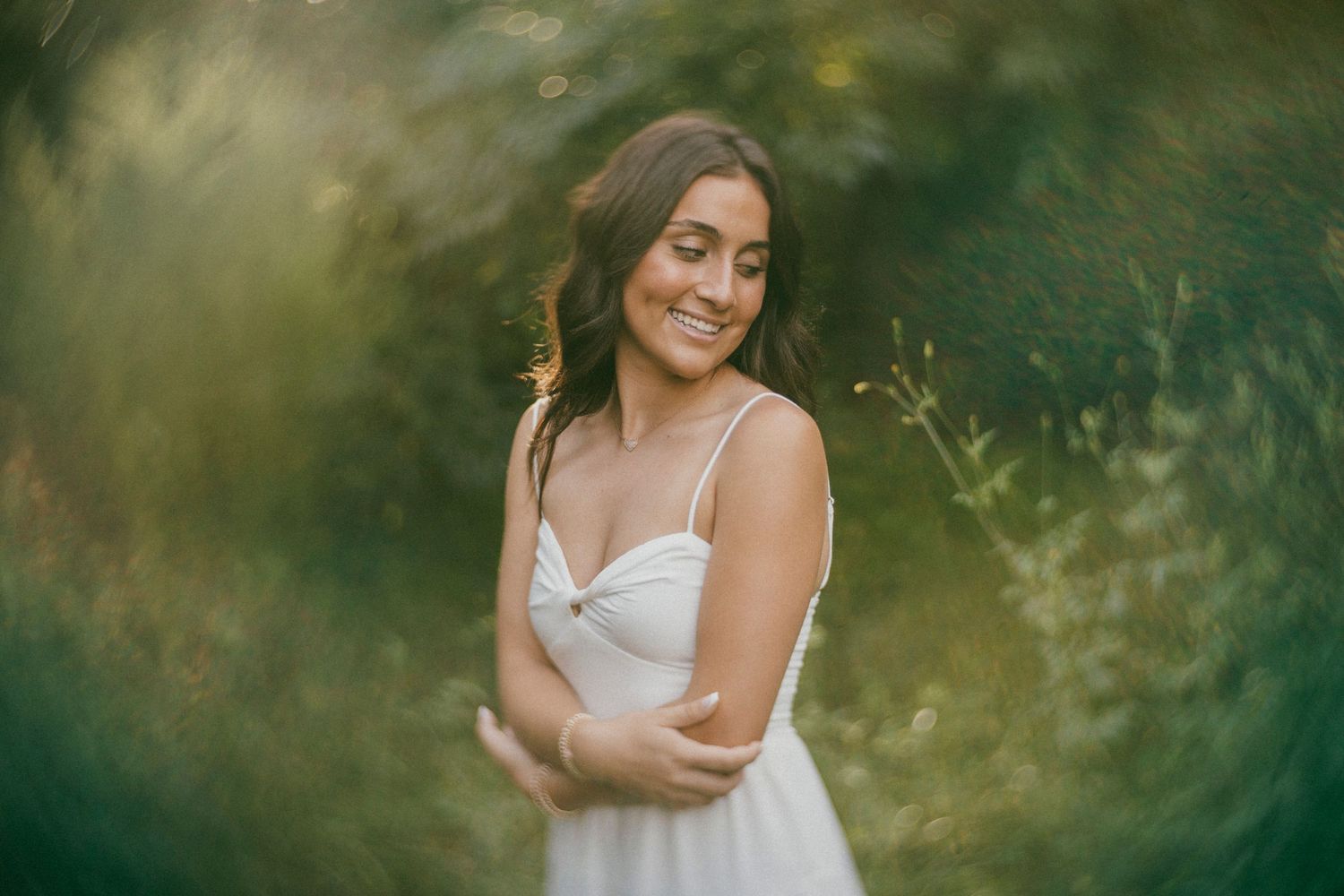 Outdoor portrait of a woman in a white dress, smiling in soft natural light with swirling bokeh and greenery in the background.
