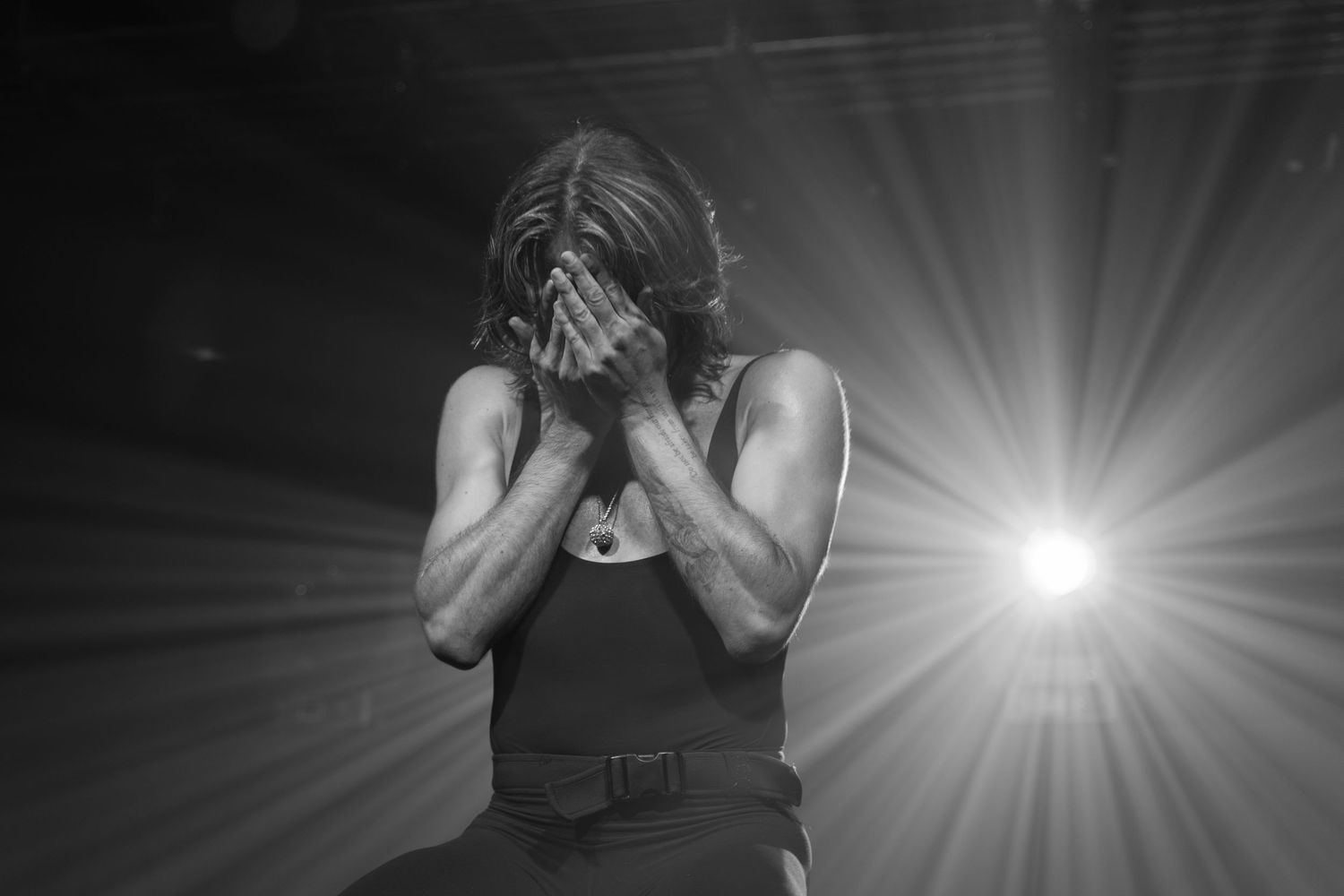 Black and white dramatic stage portrait of a performer covering their face, backlit with radiant beams of light.