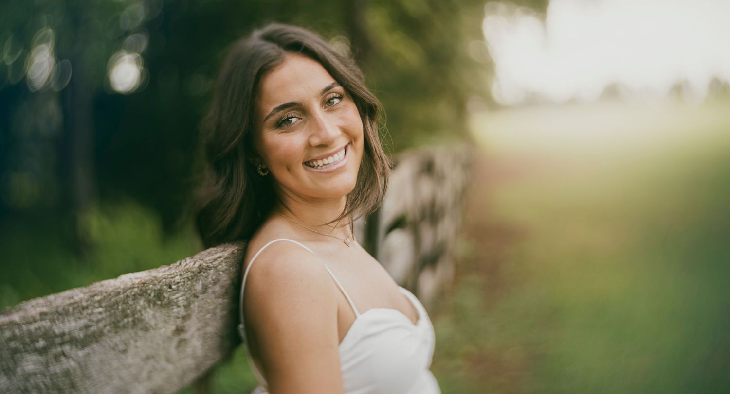 Outdoor portrait of a woman in a white dress, smiling while leaning against a weathered wooden fence with greenery in the background.