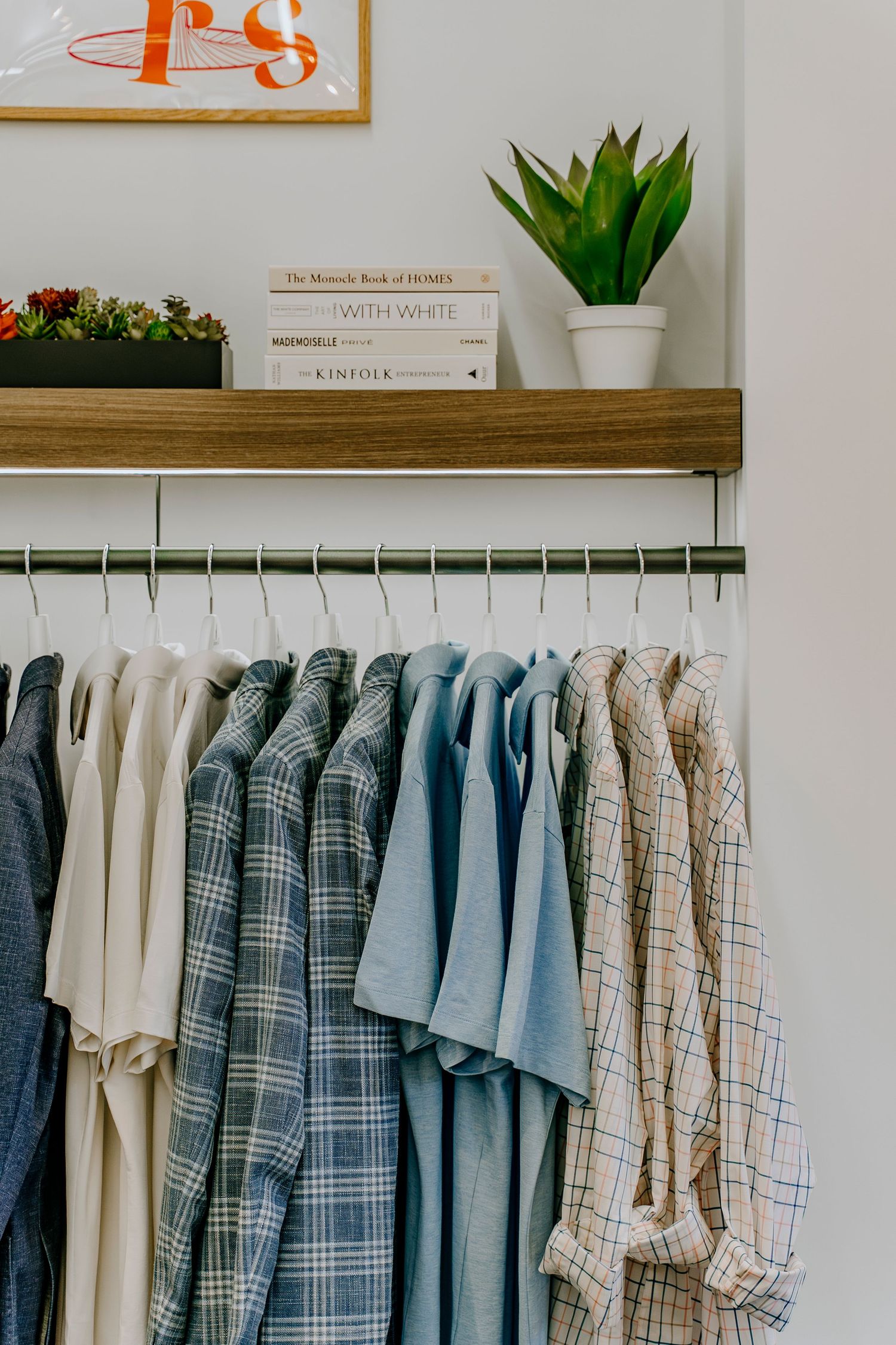 Clothing rack with neatly hung shirts in neutral and blue tones, styled under a shelf with plants and funky decor.