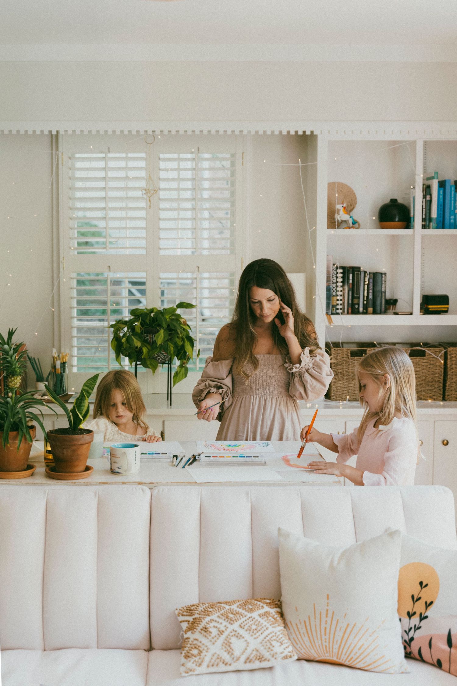 Mother helping children with arts and crafts in a naturally lit, cozy living room decorated with soft pink furniture and green plants.