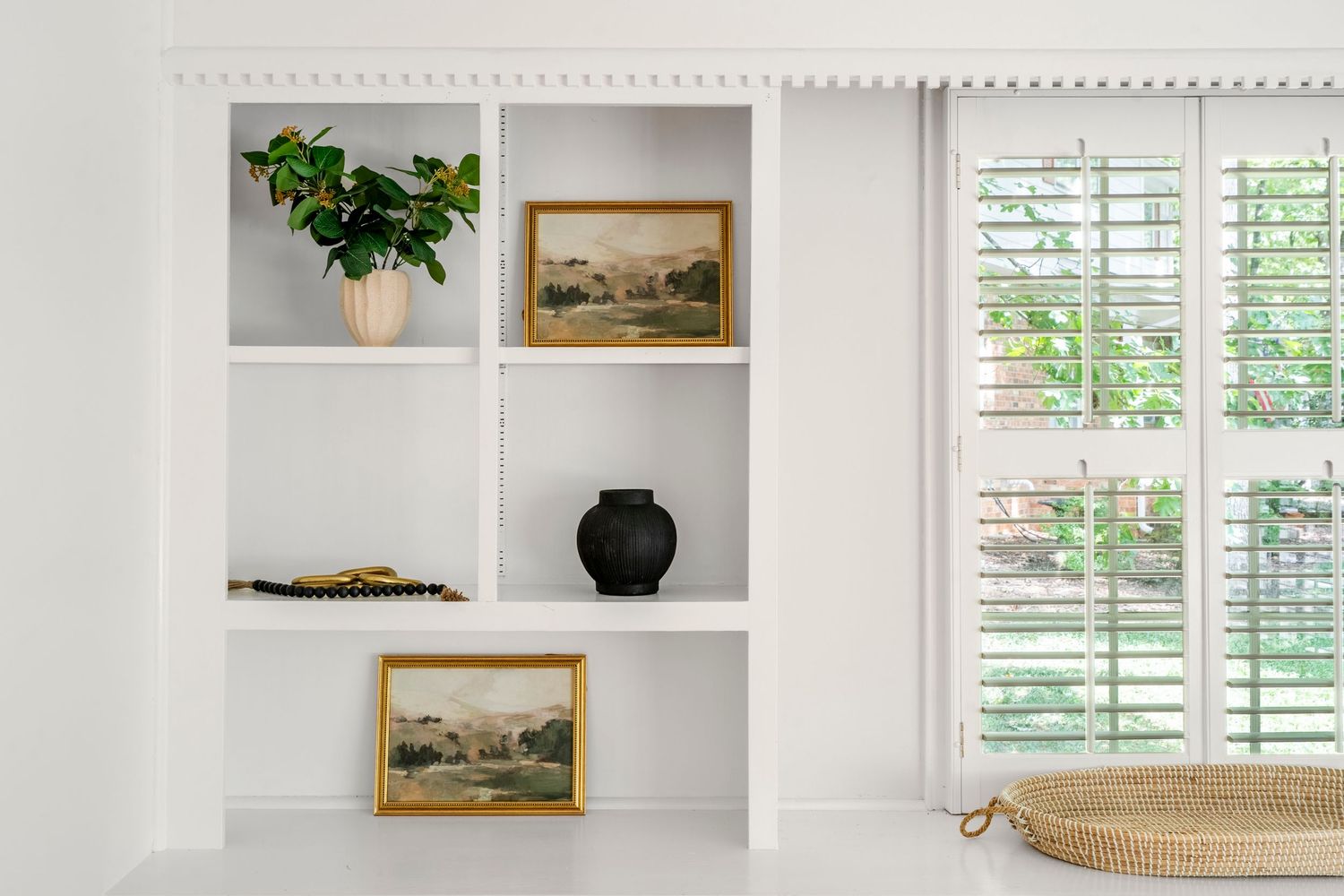 Bright interior photo of white built-in shelves styled with plants, framed art, and decorative accents beside shuttered windows.