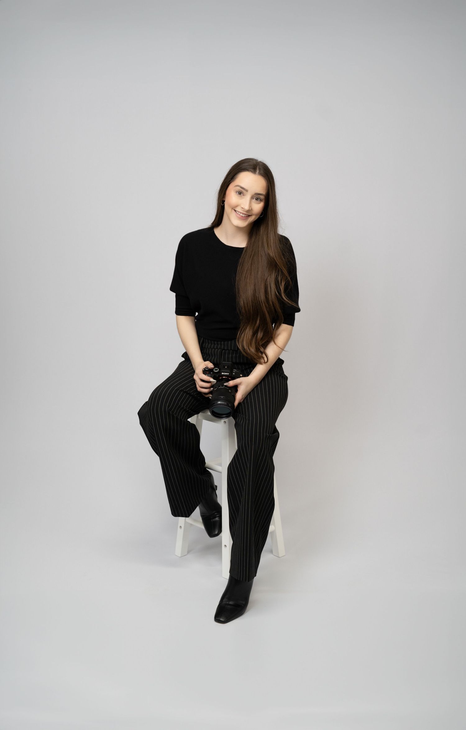 Professional female photographer (Ellie) with long brown hair smiling and holding camera against a clean white backdrop.