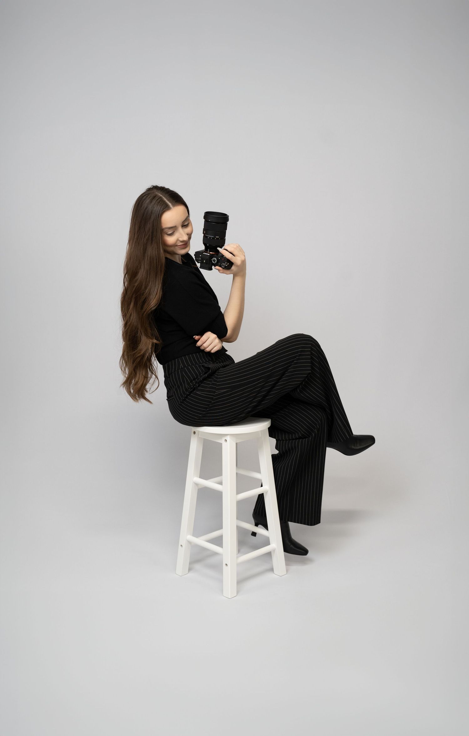 Professional woman photographer (Ellie) with brown hair posing on stool with camera in hand on clean white backdrop.