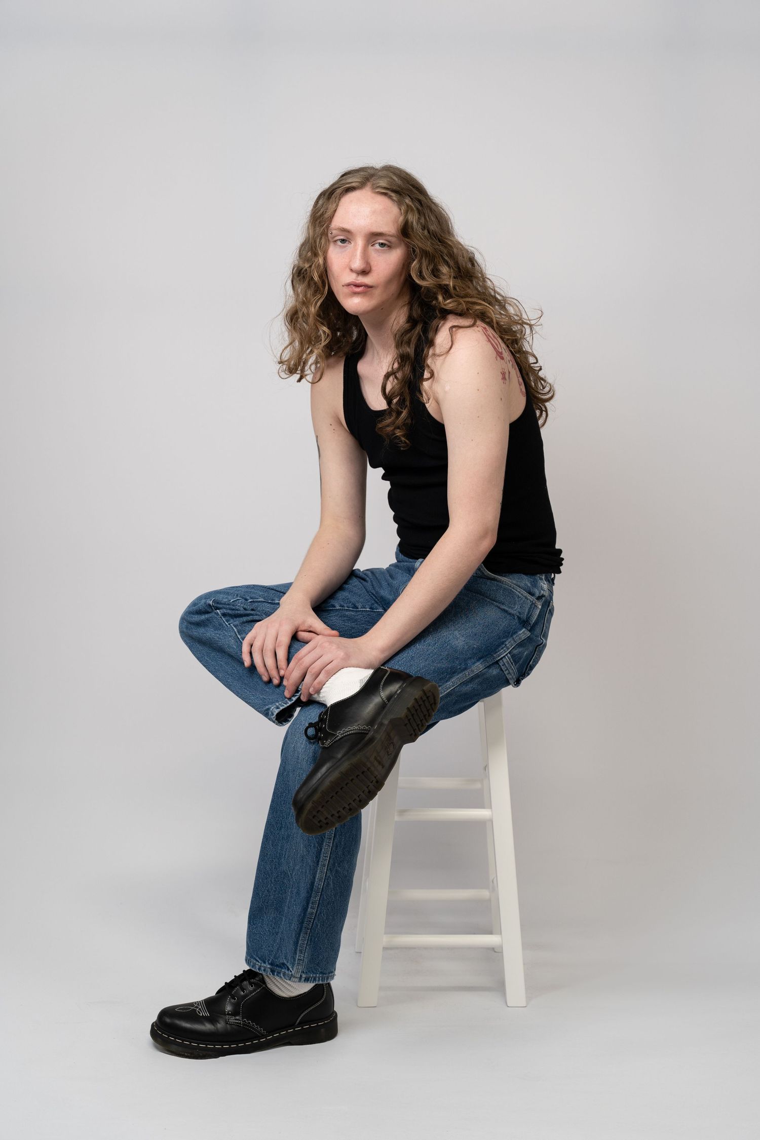 Studio portrait of a young man with long curly hair, seated on a stool in jeans and a black tank top against a gray backdrop.