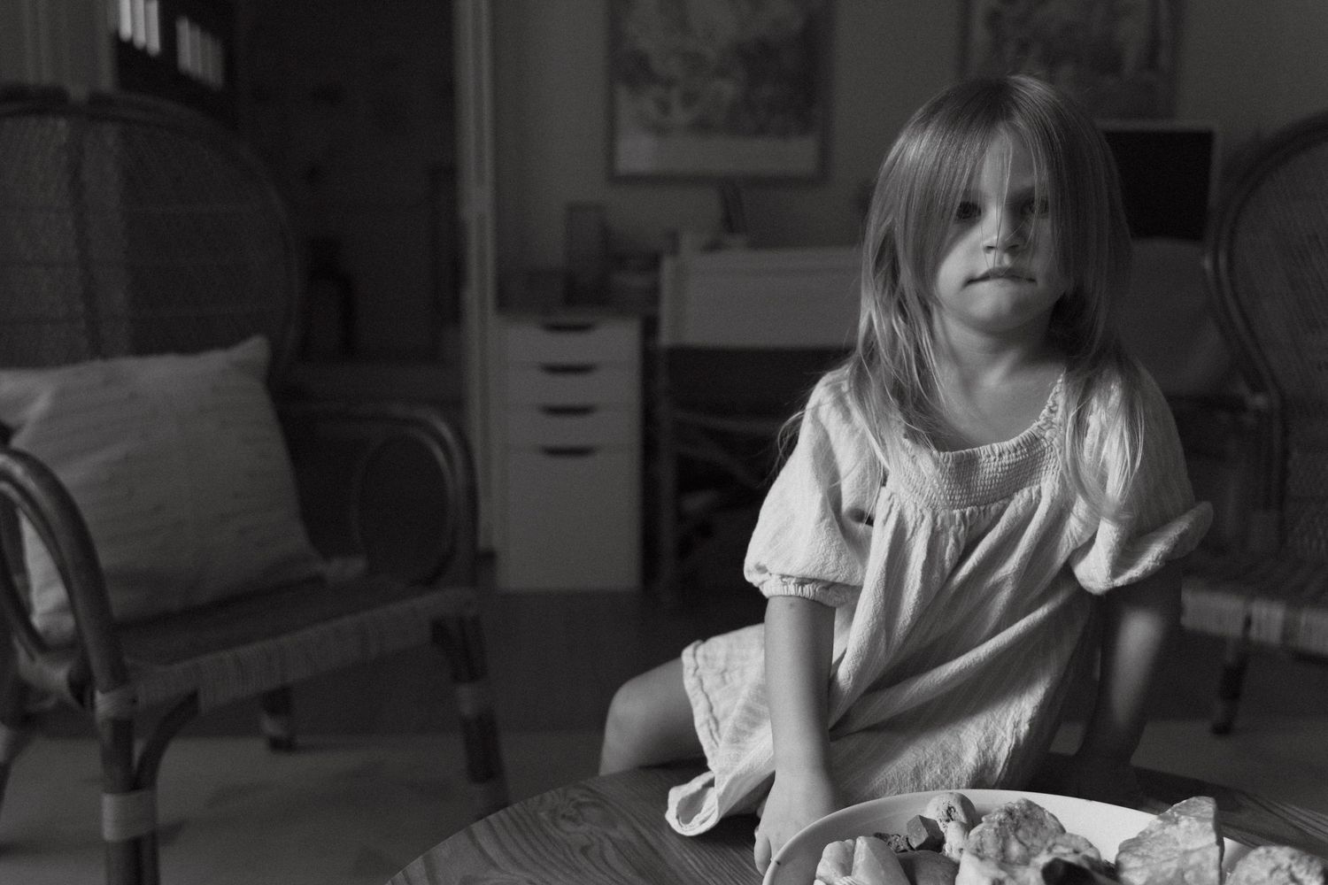 Black and white candid portrait of a young girl in a dress, seated on a coffee table indoors, framed to the right of the image.