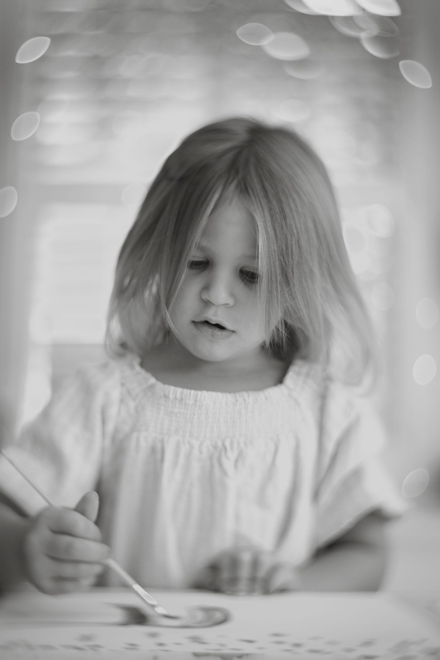 Black and white portrait of a child painting with a brush, soft natural light, and accentuated bokeh in the background.