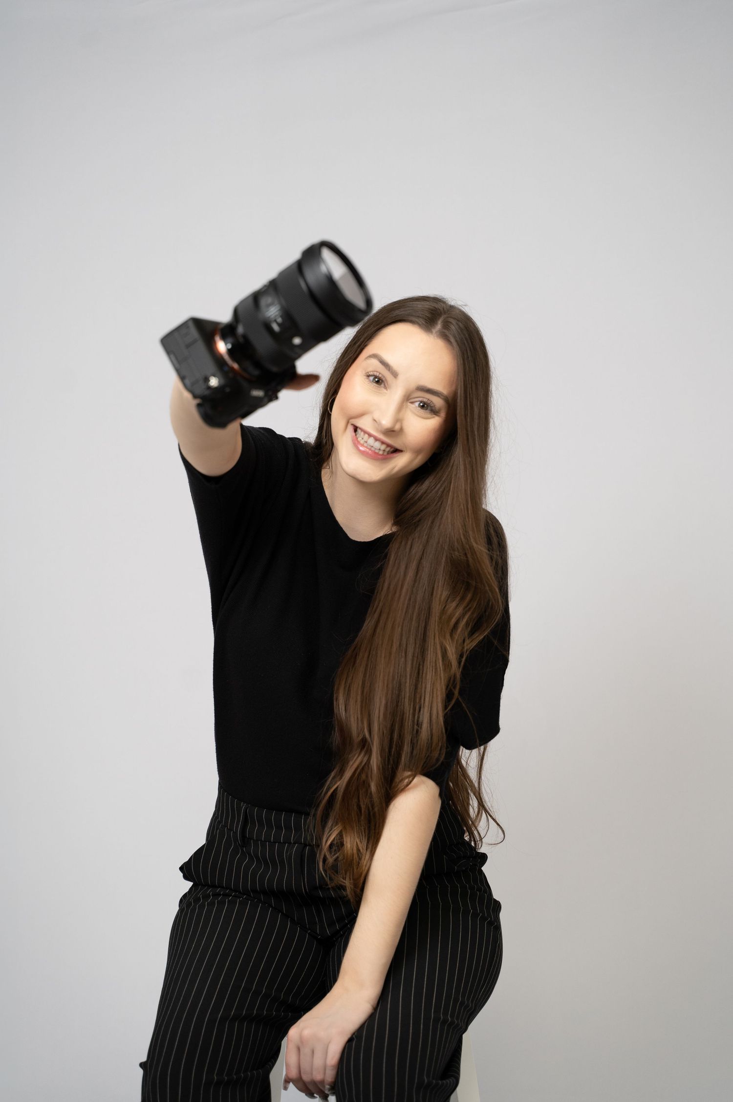 Professional female photographer (Ellie) with long brown hair smiling and holding camera against a clean white backdrop.