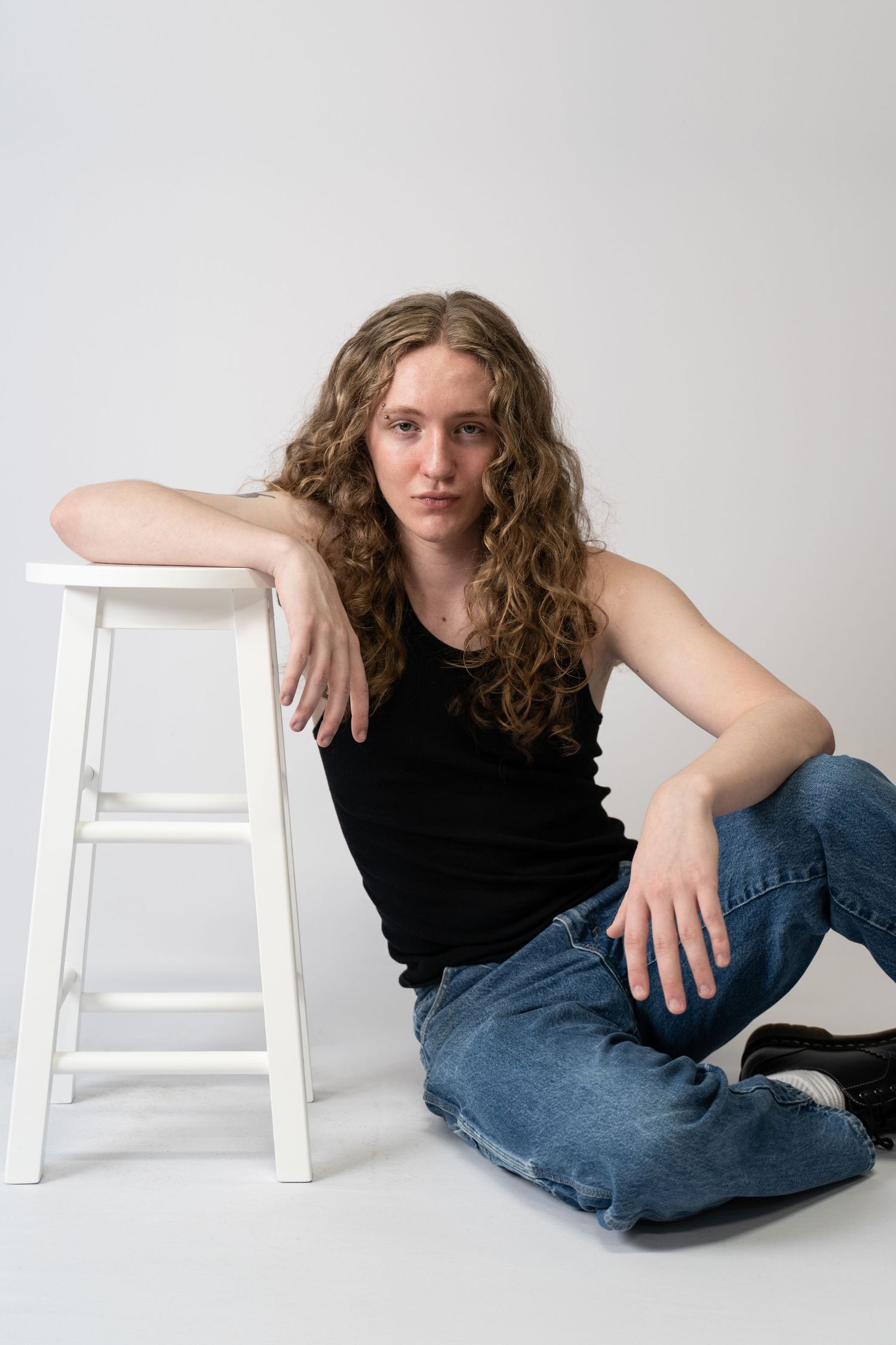 Studio portrait of a young man with long curly hair, seated next to a white stool in jeans and a black tank top against a gray backdrop.