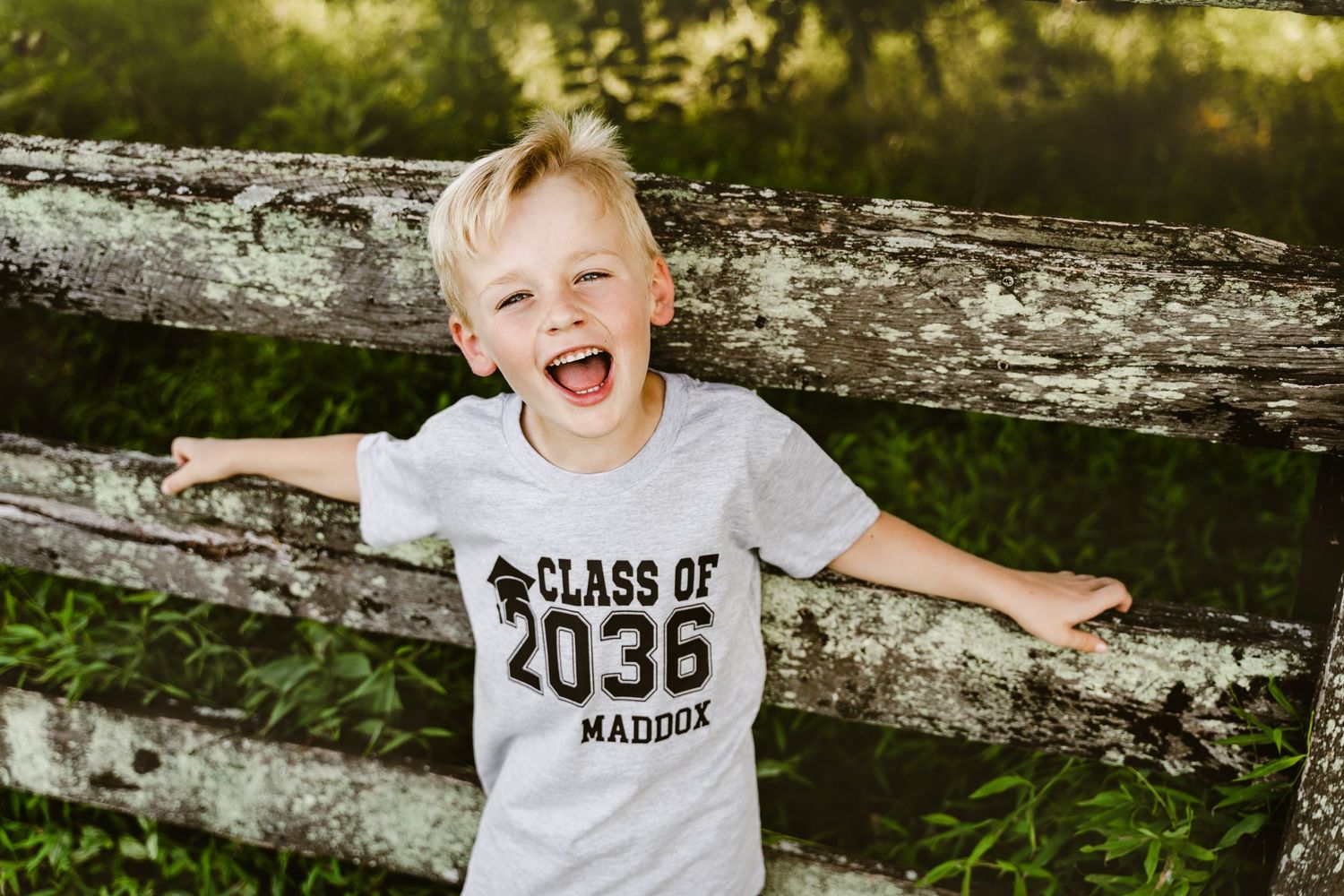 Outdoor portrait of a smiling, cheerful young boy in a “Class of 2036” shirt, leaning on a rustic wooden fence.