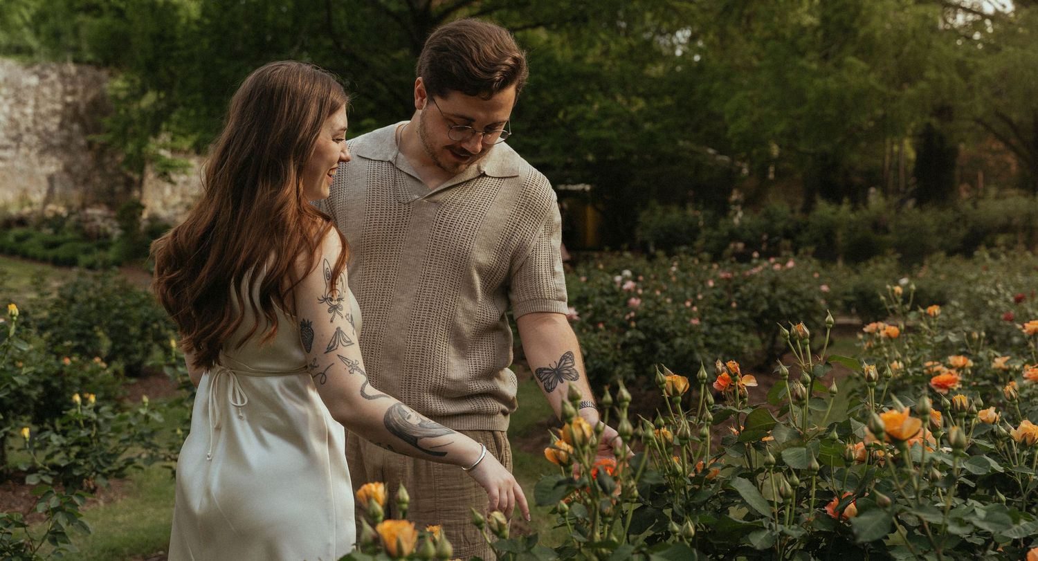 Romantic, warm-toned outdoor photo of a couple walking through a blooming garden, smiling while admiring vibrant flowers during golden hour.