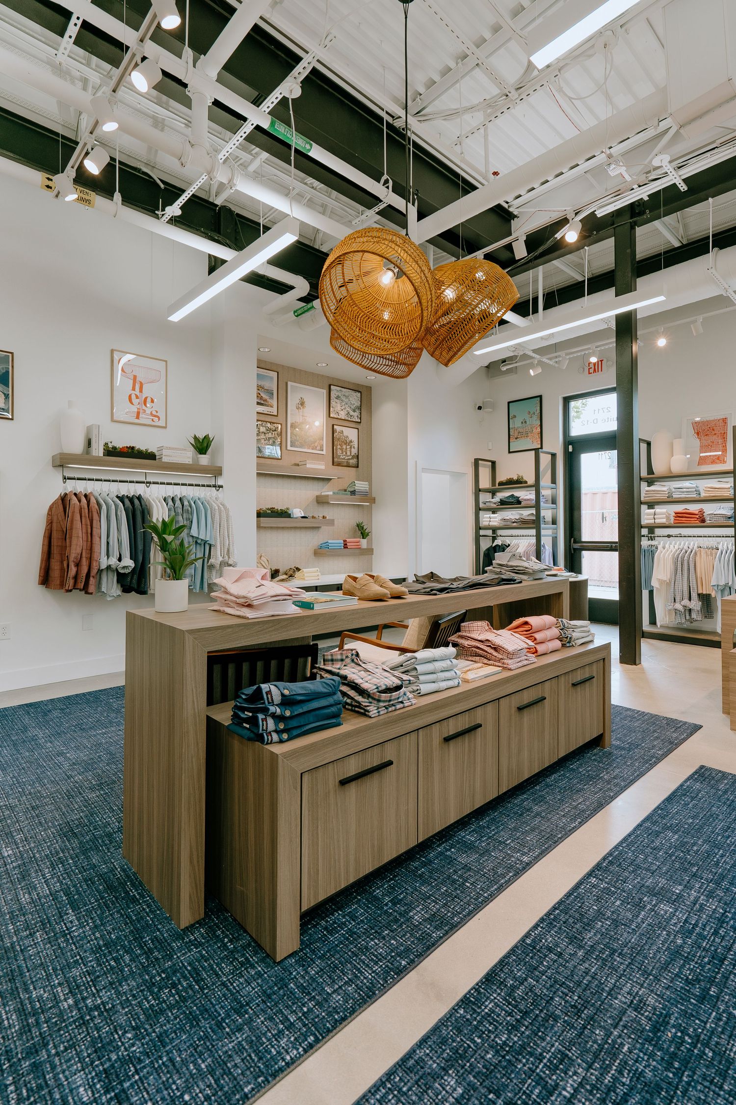 Modern Johnnie-O boutique interior with folded clothes on display tables, hanging racks, and bohemian rattan lighting fixtures.